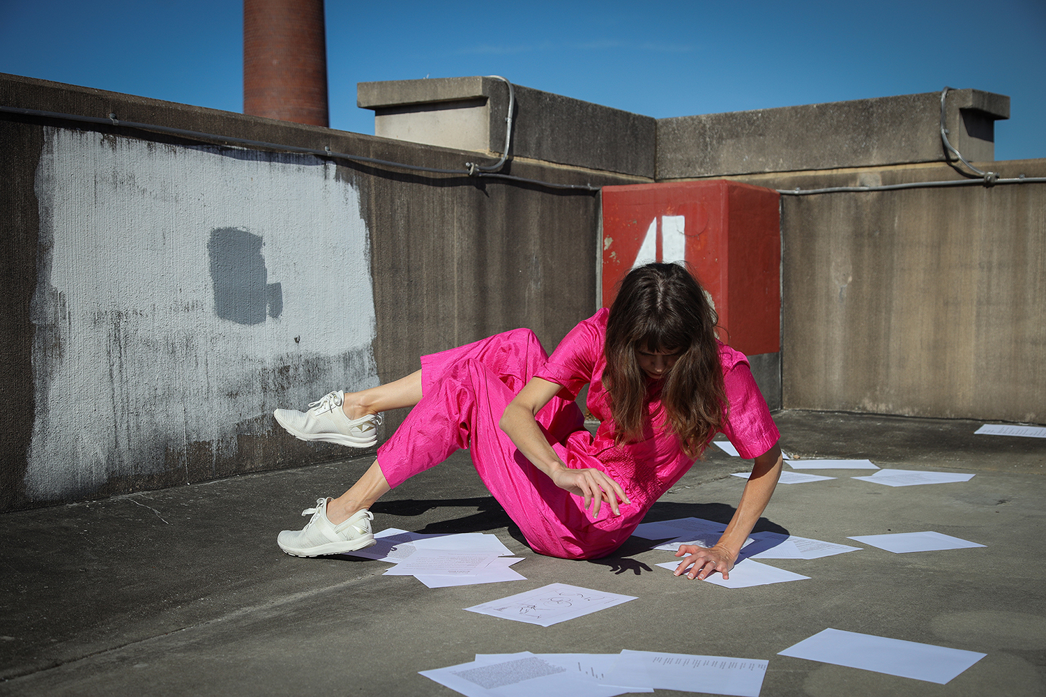 Person with long brown hair, wearing a pink jumpsuit and white sneakers, sustains a position in the corner of a concrete parking garage with papers strewn about. 