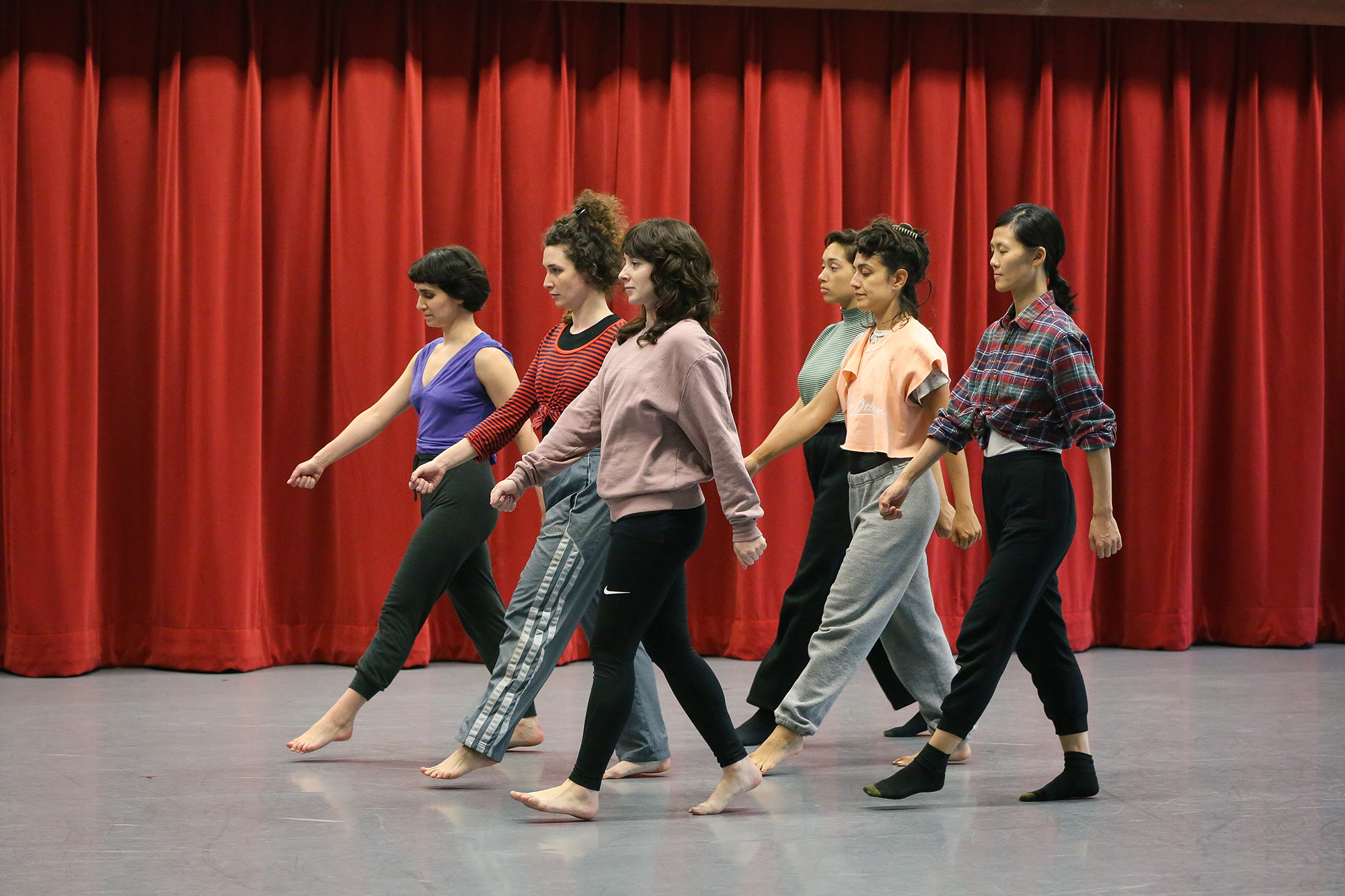 Six casually dressed people walk in unison in a two-line formation in front of a red curtain on a grey floor.