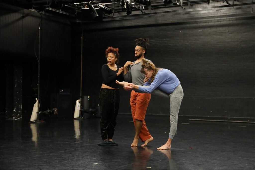One person leans in toward another standing with feet crossed. The second person is supporting another person doubled over with their leg outstretched and in their hand. They are in a theater with a black floor and scrim, with wings back, exposing the stage light equipment.