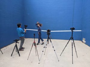 ID: One person films another dancing in front of a blue cinderblock wall. Both are behind a wall of four cameras filming at the same time.