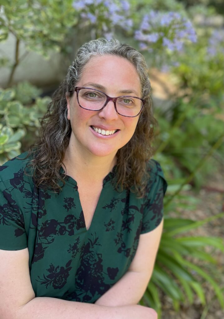 Dr. Jen Atkins is wearing glasses and has curly hair. She is dressed in a green floral shirt. She smiles, looking up towards the camera.