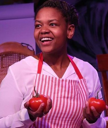 A smiling Black woman wears a white and red striped apron. She is holding one large tomato in each of her hands.