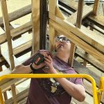 A woman uses a power tool on a structure that resembles the framing of a house.