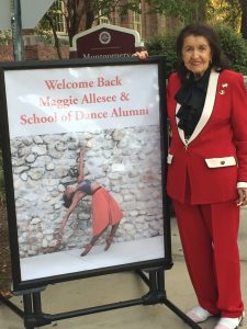 Maggie Allesee poses next to a sign that reads "Welcome back Maggie Allesee and School of Dance Alumni." She is wearing a vibrant red suit and a black blouse. 