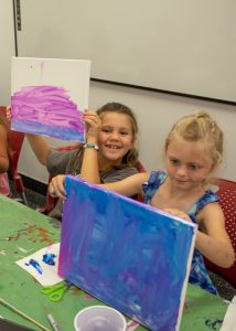 Two little girls display their abstract artwork. One on the left smiles proudly, the other has a shy expression.