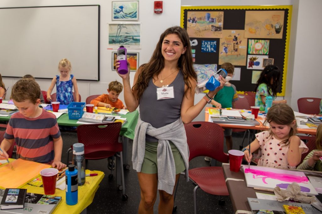 A young woman holds up bottles of paint and smiles, posing for a photo in a classroom full of children busily painting.