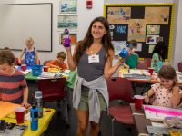 A young woman holds up bottles of paint and smiles, posing for a photo in a classroom full of children busily painting.