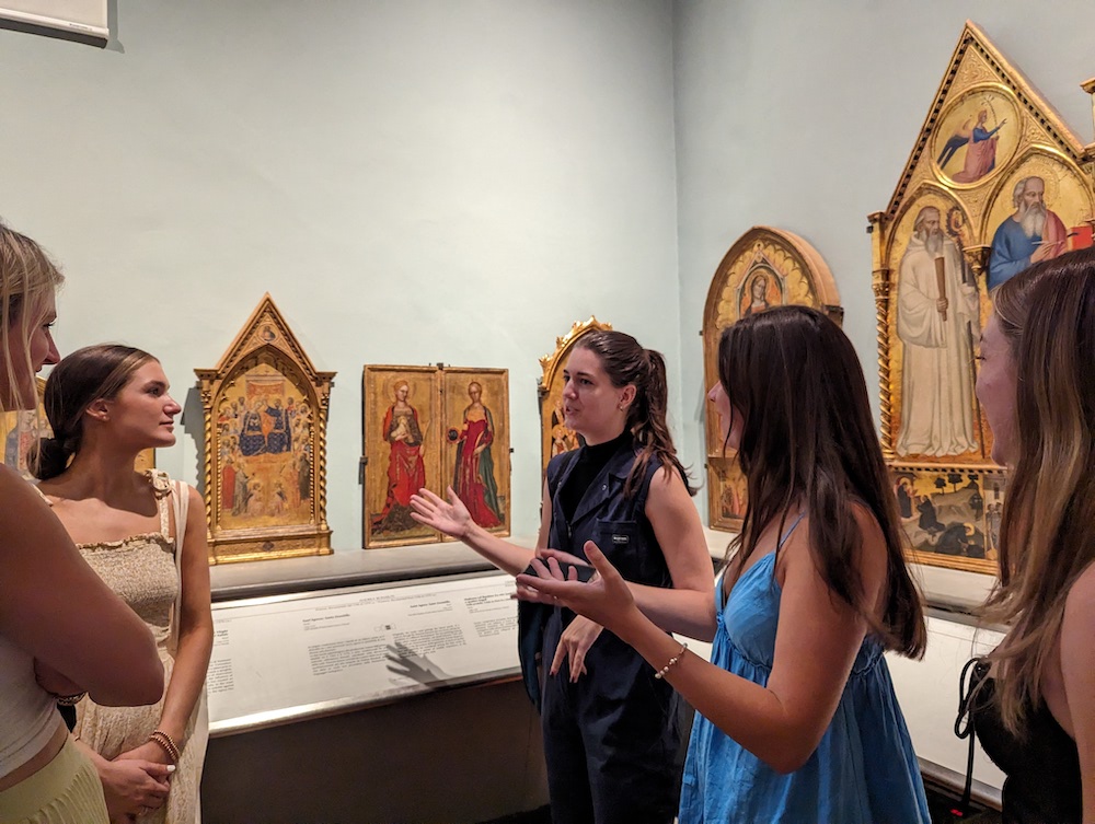 A young woman teaches in an art gallery with gilded ranaissance paintings around her. 