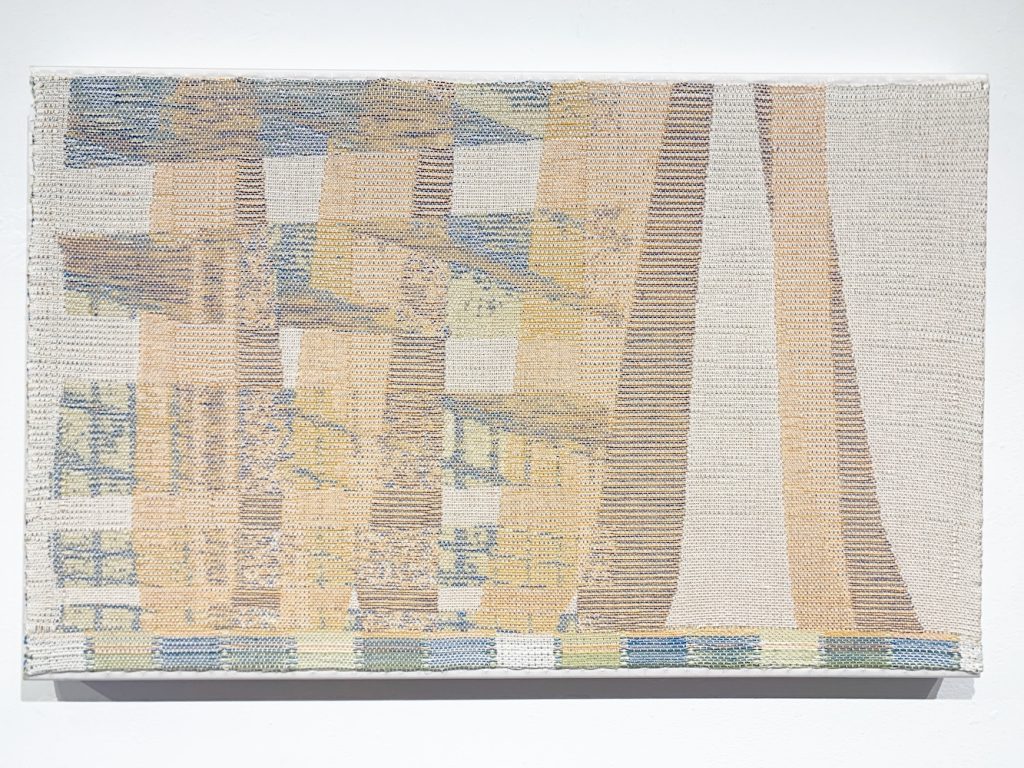 A piece of textile artwork hags in a gallery. It features an abstract pattern of neutral tones. 