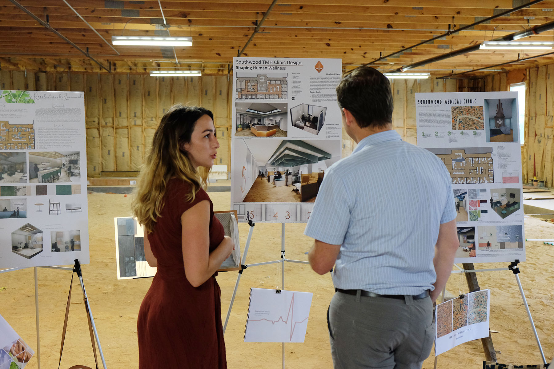 A young woman and a man in professional clothing face away from the camera, looking at a poster board with designs for a healthcare center. 