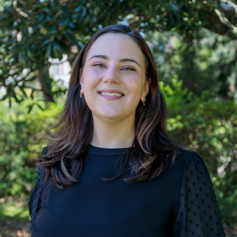 A woman smiling in a black blouse with trees and bushes in the background