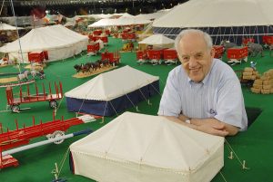 A man pops up through a hatch and smiles, surrounded by a miniature circus
