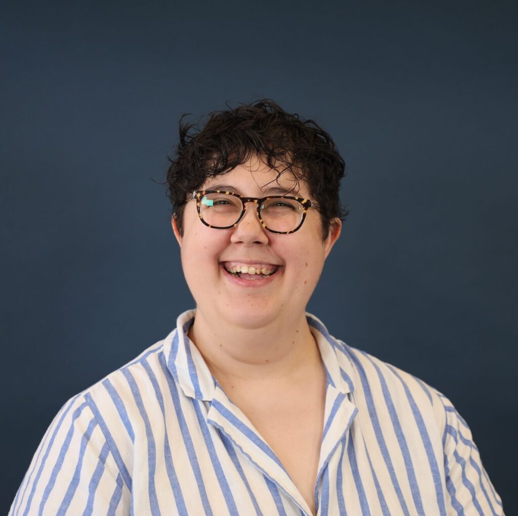 A person smiling in front of a dark blue background with short dark hair, glasses and a blue and white striped button-up shirt.