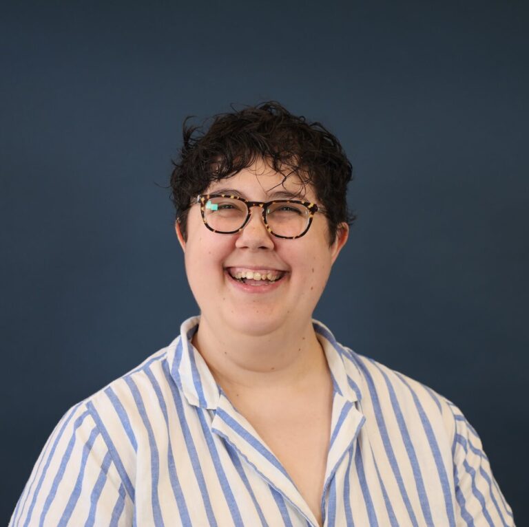 A person smiling in front of a dark blue background with short dark hair, glasses and a blue and white striped button-up shirt.