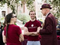 A man in a red fedora hat and matching suit talks to college students outside on a college campus.