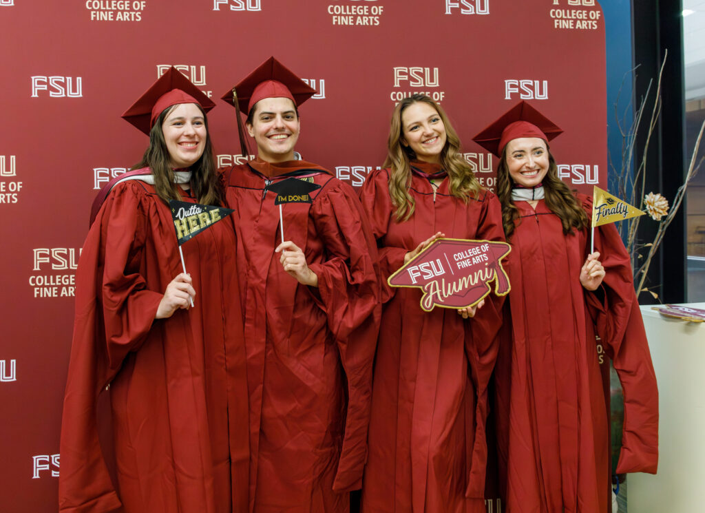 Smiling young people pose for a photo in caps and gowns, holding up celebratory flags and a sign that says "FSU College of Fine Arts Alumni"