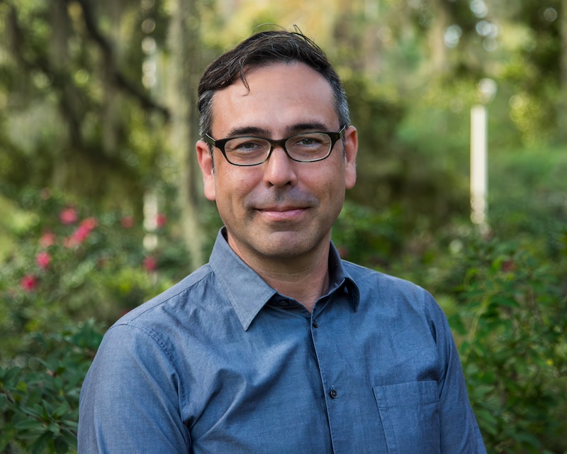 A man with glasses in a blue button-up shirt stands in front of a green, natural area.