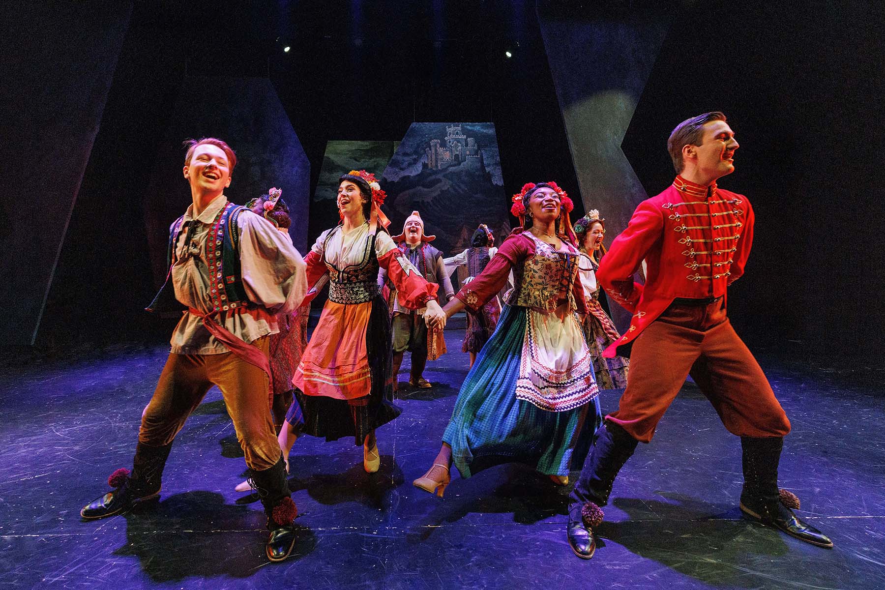 A group of colorfully costumed actors dance on stage. Behind them is a dark and moody set with a castle in the far background.
