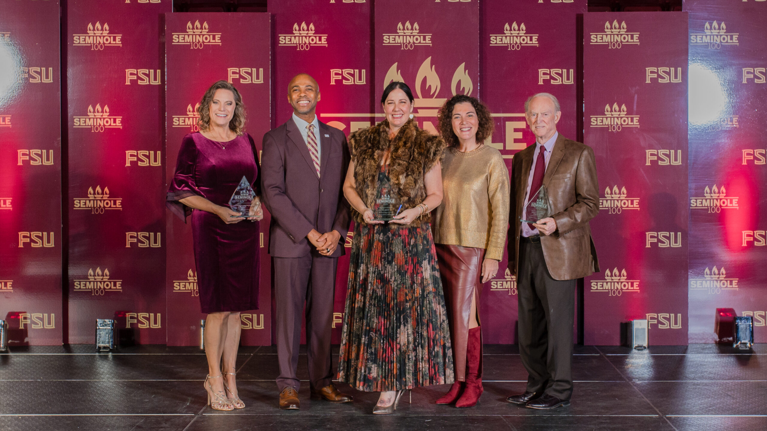 A group of people in formal attire pose for a photo, some holding glass awards. 