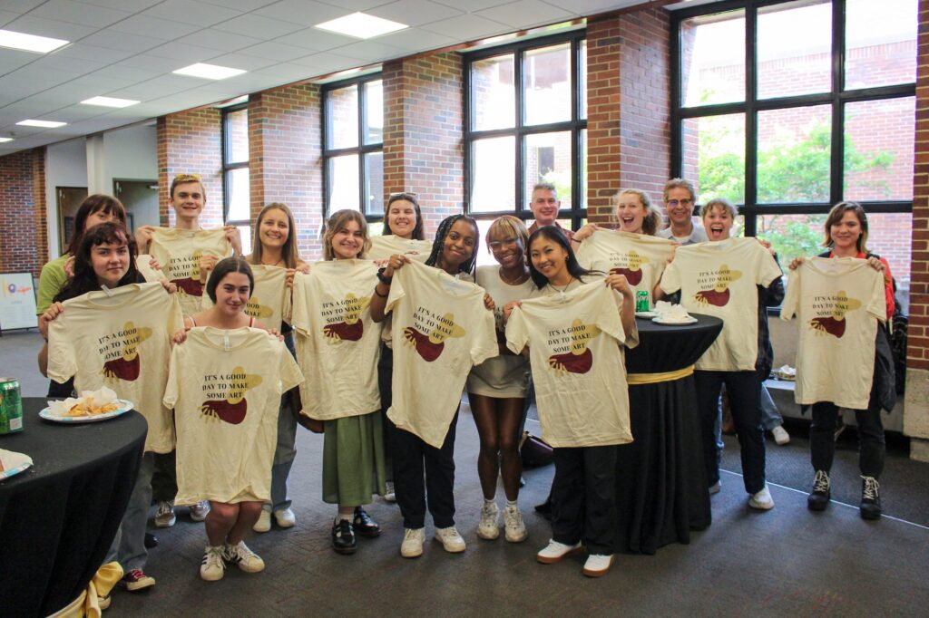 A group of college students hold up new matching T-shirts, posing for a photo in front of a wall with bright, sunny windows.