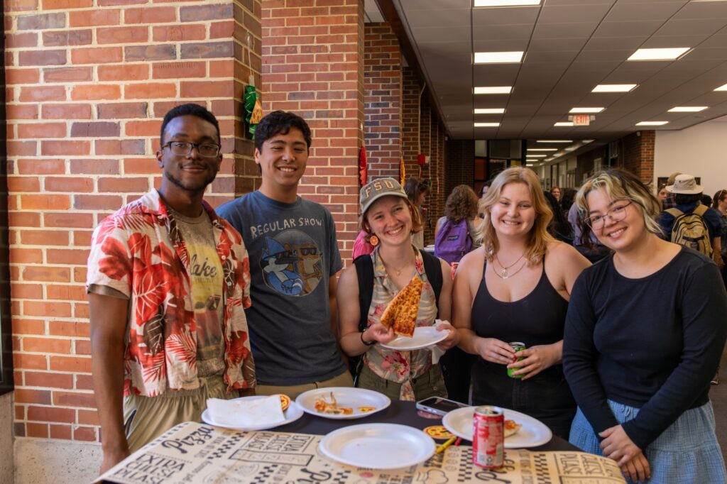 A smiling group of college students pose for a photo while eating pizza.