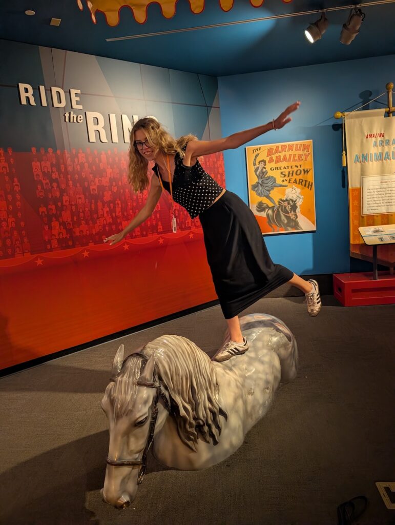 A young woman in a black dress balances on a model horse in a museum