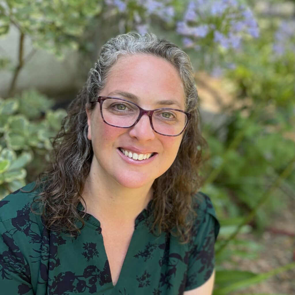 Dr. Jen Atkins is wearing glasses and has curly hair. She is dressed in a green floral shirt. She smiles, looking up towards the camera.