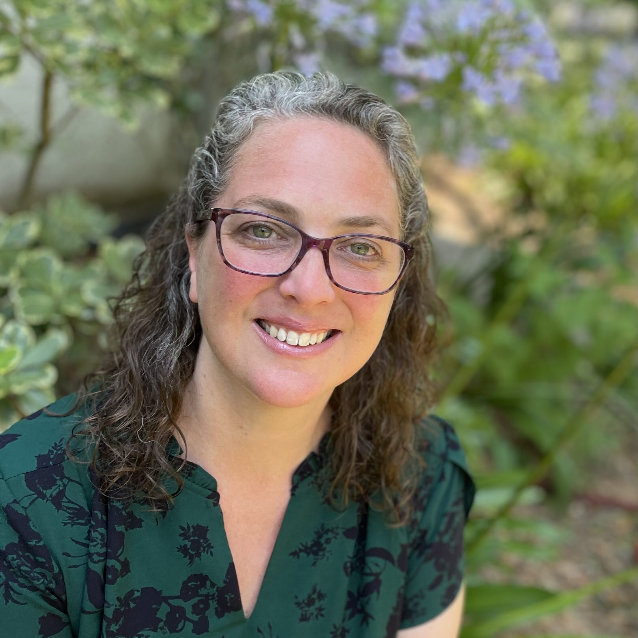 Dr. Jen Atkins is wearing glasses and has curly hair. She is dressed in a green floral shirt. She smiles, looking up towards the camera.