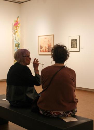 Two women sip wine while sitting on a bench in an art gallery