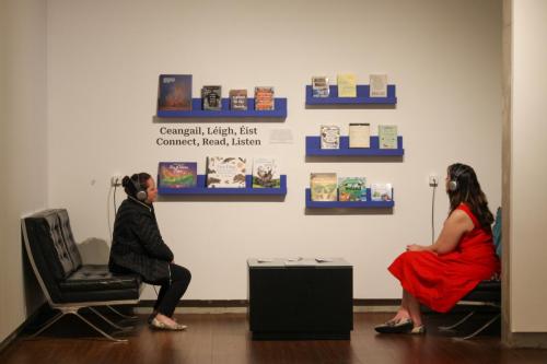 Two women wear headphones in an art gallery. They are looking at a collection of books and sitting on couches.