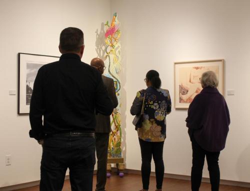 A group of people look at art in a gallery. They are drawn to a sculptural work featuring a pink bird on top.